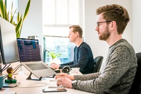 Two colleagues working side by side at bright desks with large monitors